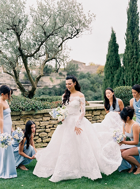 Bridal party portrait of a bride with bridesmaids as they adjust her long lace train, holding blue and white bouquets on a garden lawn by a stone wall