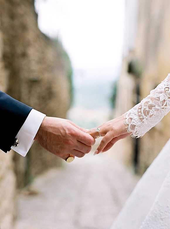Wedding ring close-up of couple holding hands, engagement ring on finger beside lace sleeve and groom cufflinks on a stone walkway with greenery