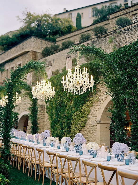 Reception tablescape for an outdoor wedding reception with a long banquet table, blue vases, hydrangeas, candles, and chandeliers in an ivy stone courtyard