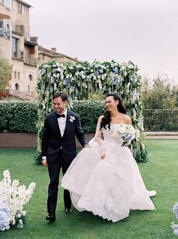 Recessional moment as bride and groom walking, smiling under a floral arch; she carries a blue hydrangea bouquet on a garden lawn