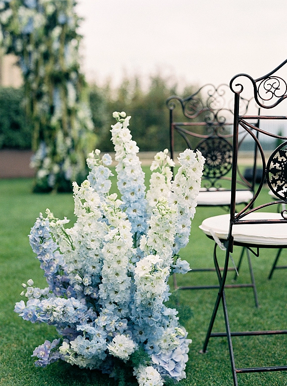 Ceremony aisle florals with blue and white flower clusters line wrought iron chairs with cushions on a grassy outdoor field under trees