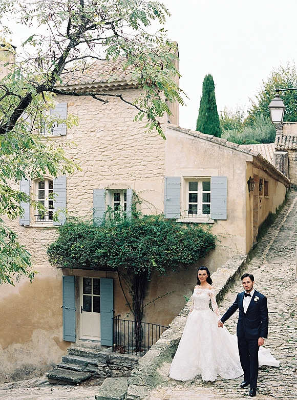 Couple portrait of bride and groom holding hands, her lace sleeve wedding dress and his tuxedo on a cobblestone street by blue shutters