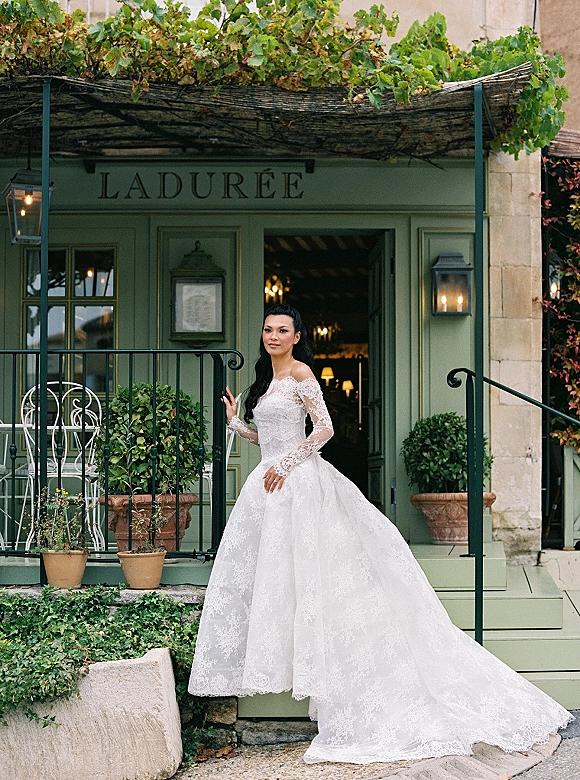 Bridal portrait of a bride in an off the shoulder lace wedding dress with long sleeves, posed by a green storefront with ivy and lanterns