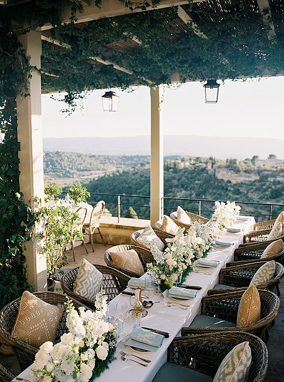 Reception tablescape with an outdoor reception table set with white florals, greenery garland, and wicker chairs under a pergola with mountain views