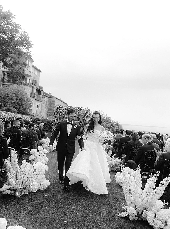 Wedding recessional as bride and groom walk hand in hand down a flower-lined aisle, guests applauding by a stone estate with water view