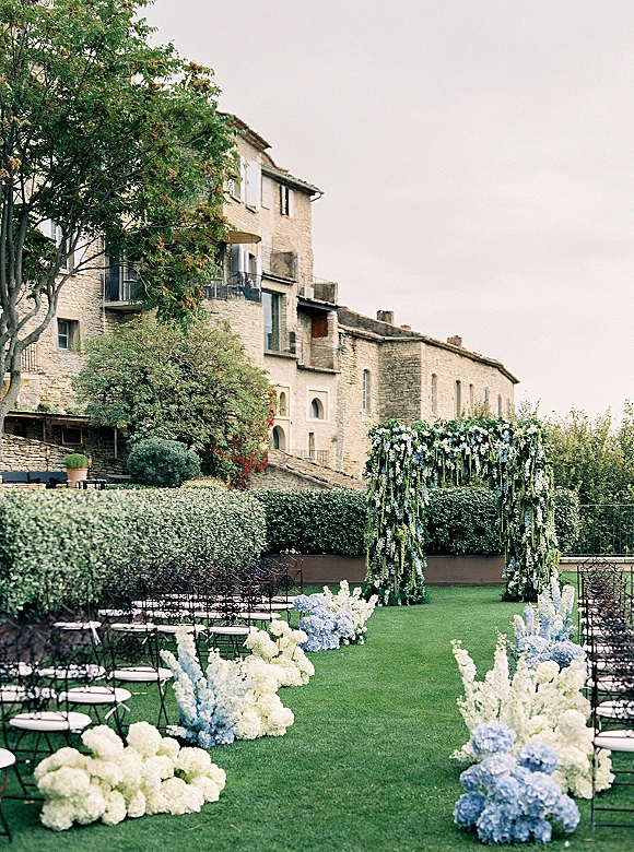 Ceremony setup with a floral arch and hydrangea aisle flowers lining the lawn aisle, metal chairs facing a stone building and hedges