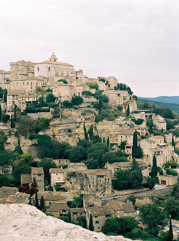 Destination wedding location overlooking a historic stone village with church and terracotta roofs, cypress trees, and cloudy hillside valley views