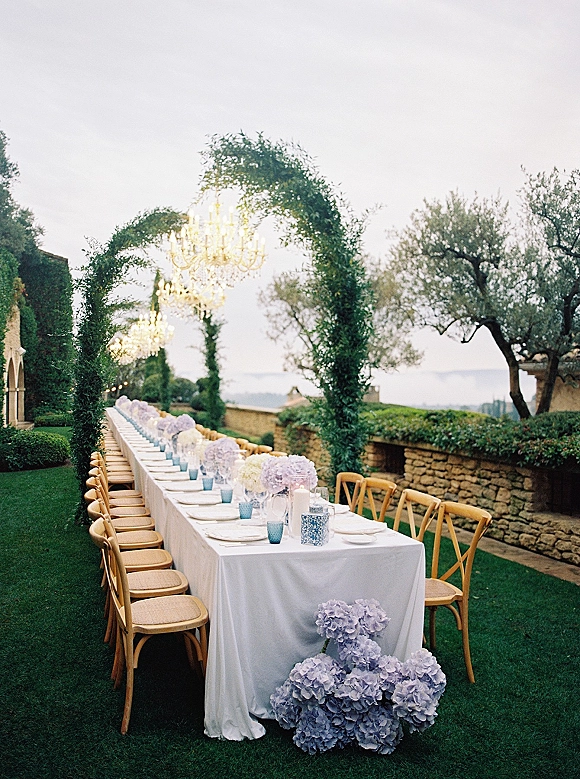 Outdoor reception table set with white linen, blue water glasses, and hydrangea centerpieces beneath chandeliers on a villa garden lawn