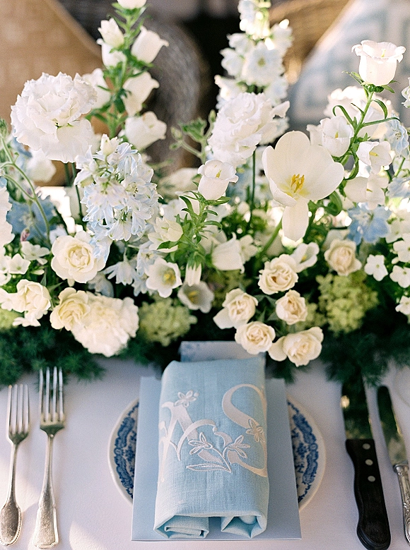Reception tablescape with wedding place setting, blue menu card, embroidered light blue napkin, blue-and-white plate, silver flatware, and white florals.