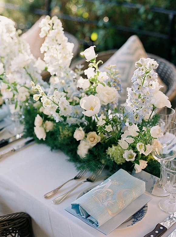 Reception tablescape with wedding table centerpiece of white blooms and greenery runner, blue napkin and menu card, glassware on patio table