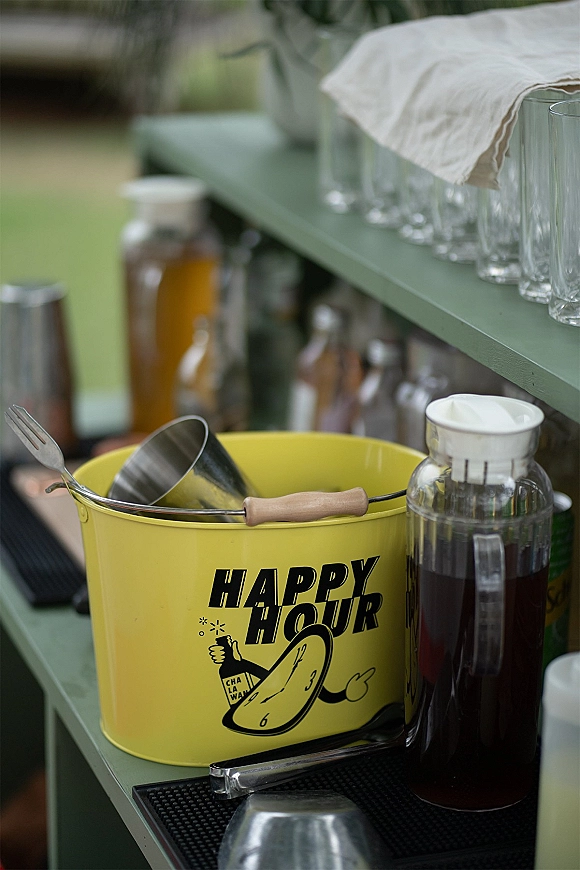 Wedding bar setup with an outdoor wedding bar on a lawn, featuring a yellow drink bucket, glassware, carafes, and bar tools on shelves