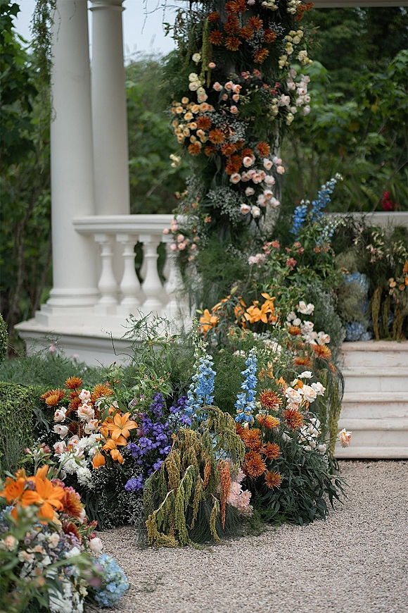 Ceremony altar florals framing a wedding floral arch with roses, lilies, and delphinium on white porch columns and steps in a garden setting