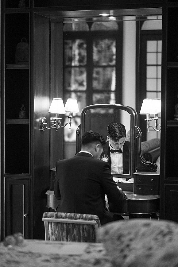 Groom getting ready in a tuxedo, adjusting his bow tie at a vanity mirror in a dark wood room with warm sconces and shelves