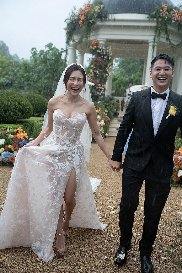 Wedding recessional as bride and groom hold hands, laughing in rain on a gravel garden aisle under a floral arch with rose petals