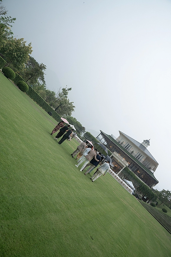 Wedding guest arrival with umbrellas as guests in raincoats and suits walk across a lawn toward a pavilion under an overcast sky