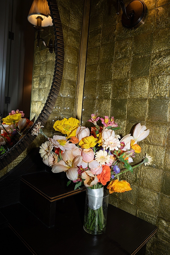 Wedding bouquet of mixed colorful flowers in a glass vase with ribbon wrap, styled on a black console by a round mirror and gold wall