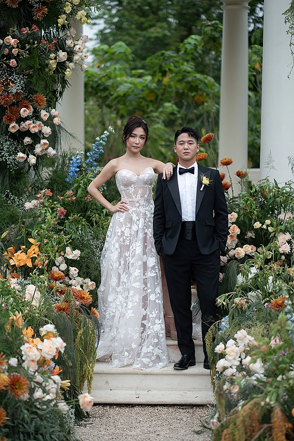 Couple portrait of bride and groom posing on stone steps, her hand on his shoulder, beneath a floral arch with roses and greenery outdoors