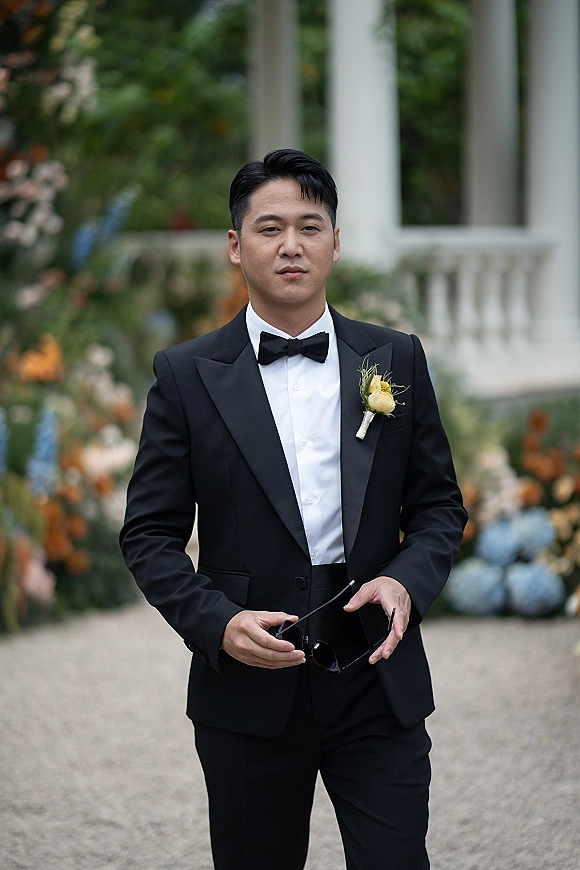 Groom portrait in a black tuxedo and bow tie, holding sunglasses with boutonniere, standing by white columns in garden greenery