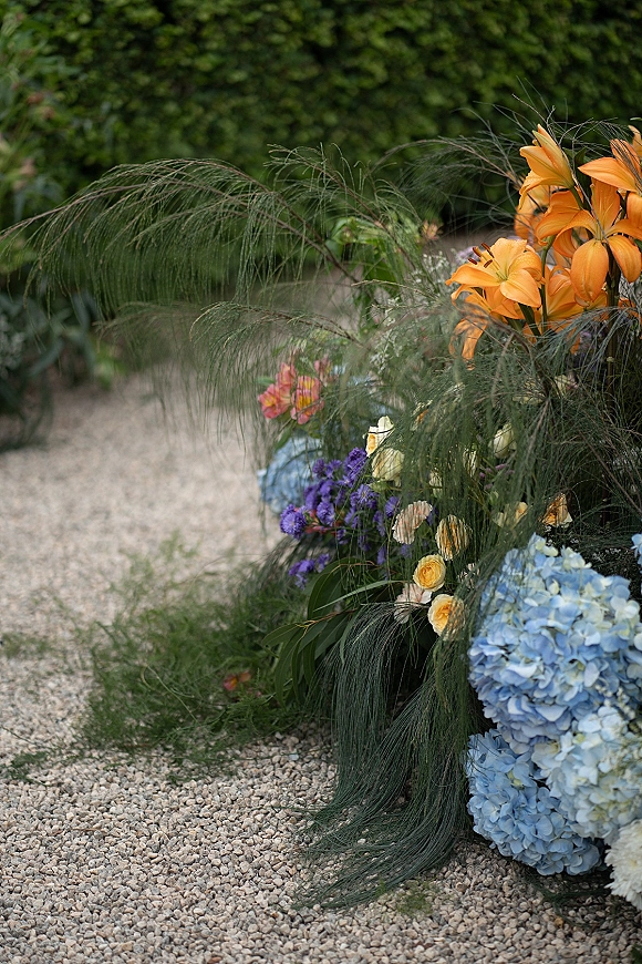 Wedding floral arrangement with ceremony aisle flowers, featuring orange lilies and blue hydrangeas beside a gravel garden path hedge