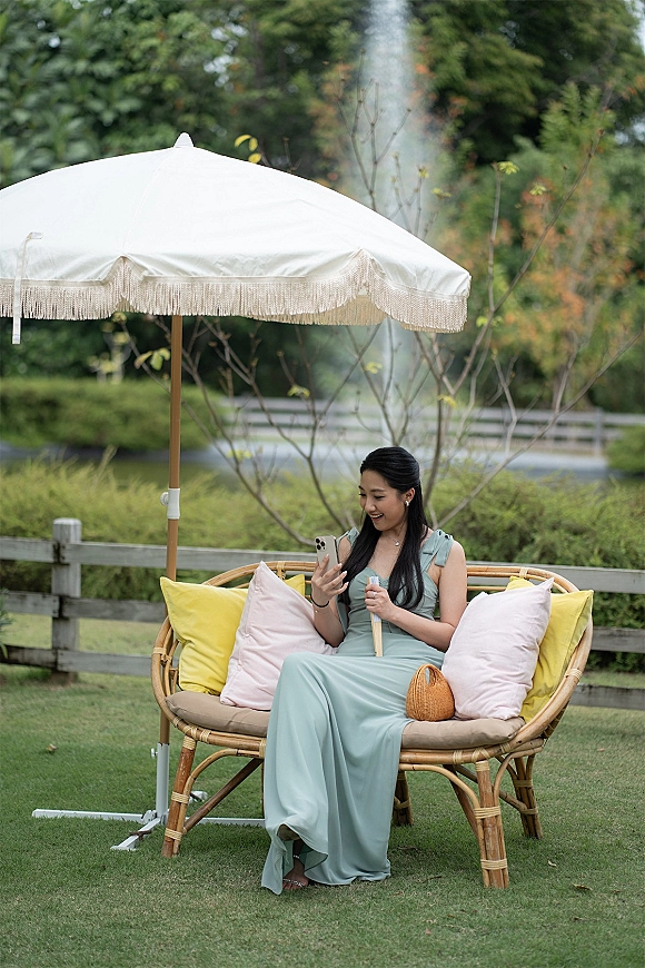 Bridesmaid portrait in a sage dress, seated on a wicker bench holding a phone, with umbrella and garden lawn backdrop