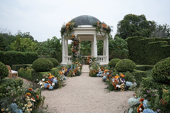 Ceremony aisle decor with blue hydrangeas and garden roses lining a gravel walkway to a white gazebo in a formal garden