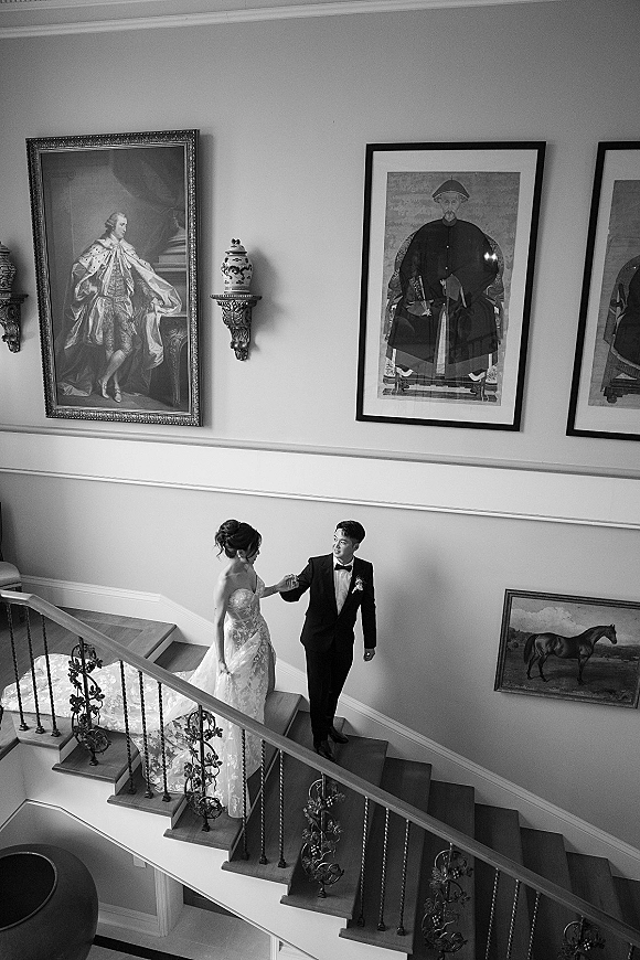 Couple portrait of bride and groom on staircase, holding hands as she lifts her dress train beside framed artwork and railing