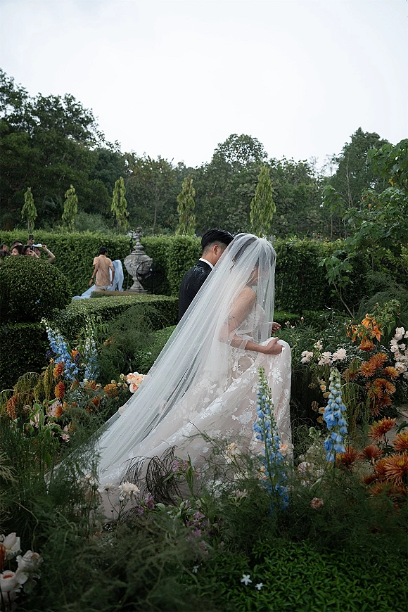 Couple portrait of bride and groom embracing, her long veil and lace dress flowing amid colorful garden flowers and hedges