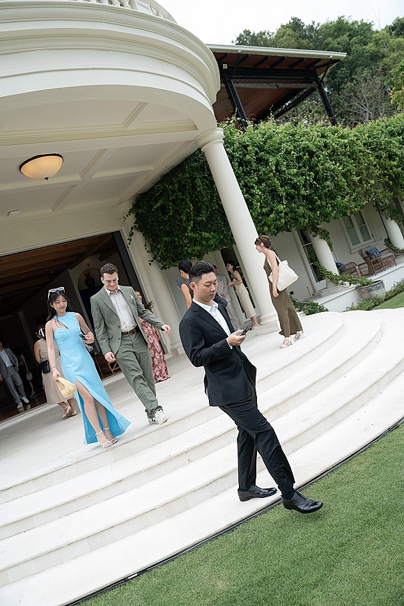 Wedding guests in formal attire walking up white veranda steps with columns, ivy-covered wall, and one blue satin dress accent