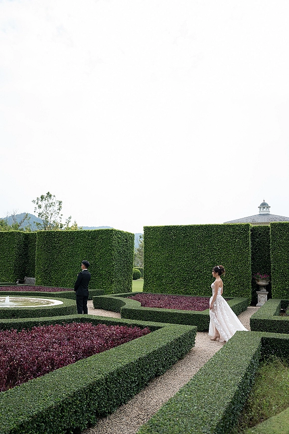 Wedding first look as bride in a strapless ballgown approaches groom in a black tuxedo on a gravel path by hedges and fountain