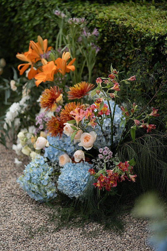 Wedding floral arrangement with orange lilies and blue hydrangeas, peach roses, and greenery beside a garden hedge on a gravel path