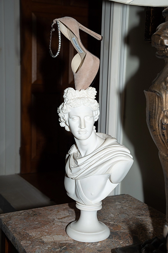 Bridal shoes in a wedding shoes flat lay, nude high heels with rhinestone anklet on a marble tabletop beside a classical bust statue