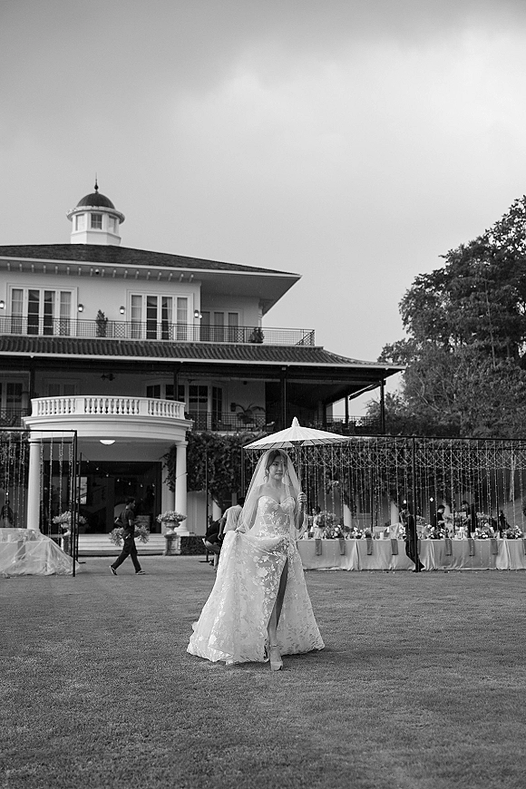 Bridal portrait of a bride holding parasol, veil flowing over a lace wedding dress on a lawn before a historic mansion with string lights