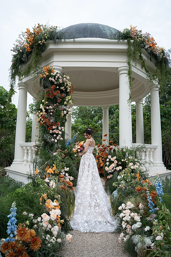 Bridal portrait of a bride holding bouquet in a strapless lace wedding dress with long train beneath a white gazebo with colorful florals