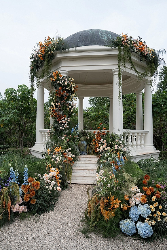 Ceremony altar decor with wedding gazebo flowers, lush greenery garlands and orange blooms on a white gazebo along stone steps in a garden setting