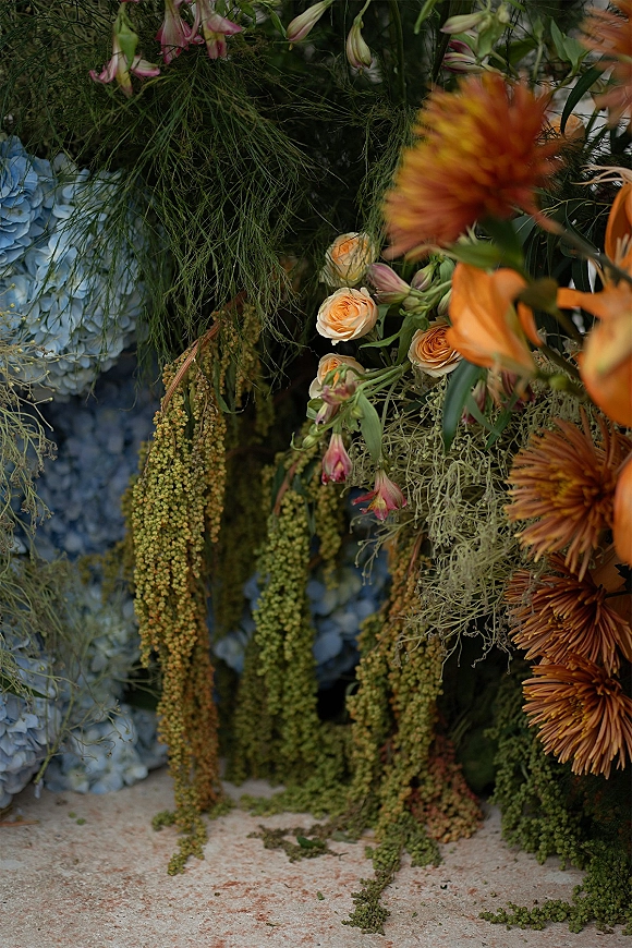 Wedding florals in a ceremony floral installation with orange chrysanthemums, peach roses, blue hydrangea, and hanging amaranthus on stone