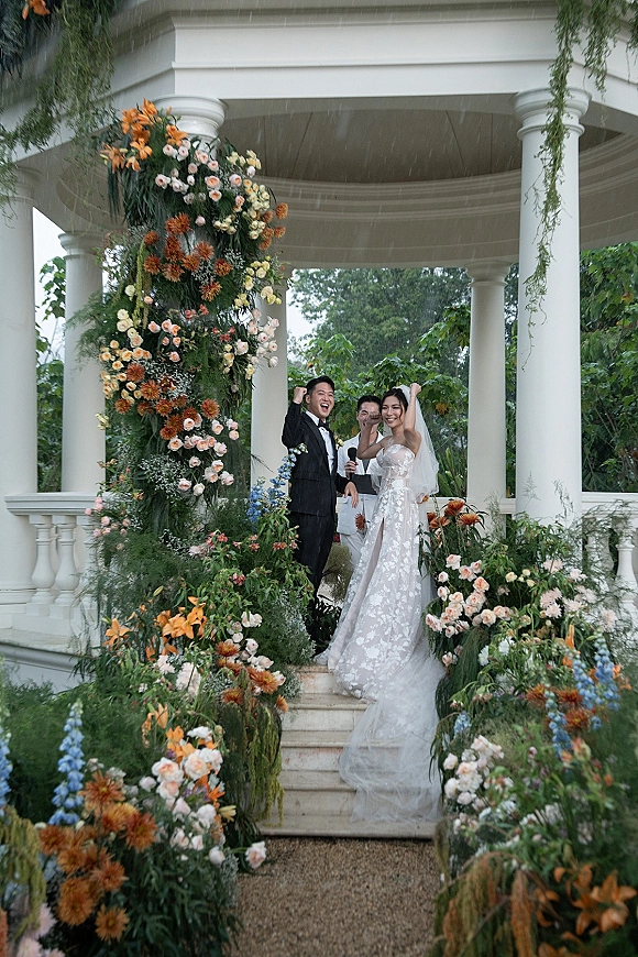 Ceremony moment as bride and groom cheer under a floral arch at a white gazebo in the rain, veil lifted beside tuxedoed groom