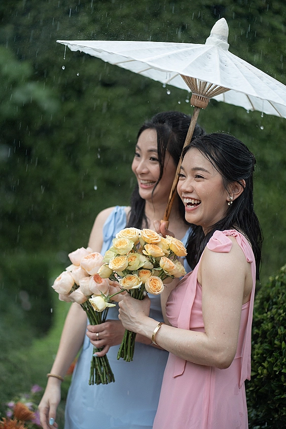 Bridesmaid portrait of two bridesmaids in rain under a white umbrella, holding rose bouquets in pink and blue dresses by garden hedge