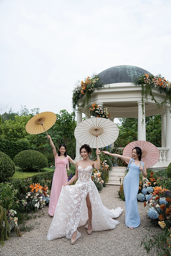 Bridal party portrait of bride with bridesmaids holding parasols in pastel dresses, posing by a floral gazebo in a garden walkway