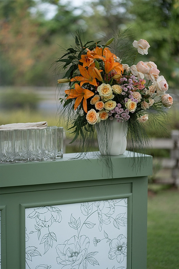 Wedding floral arrangement with orange lily centerpiece of blush and peach roses in a white ceramic vase on a painted bar cart in a garden