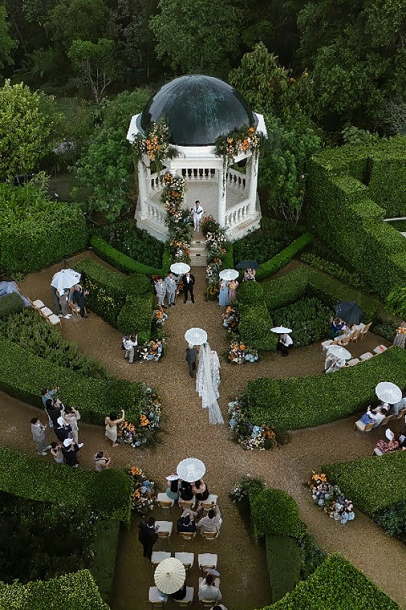 Outdoor ceremony setup with garden wedding ceremony aisle, white chairs and guest umbrellas around a floral arch and gazebo in formal hedges