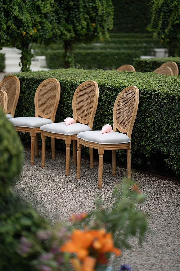 Ceremony seating with outdoor ceremony chairs, cane-back wood chairs and white cushions, pink programs on seats along a gravel aisle in a hedge garden
