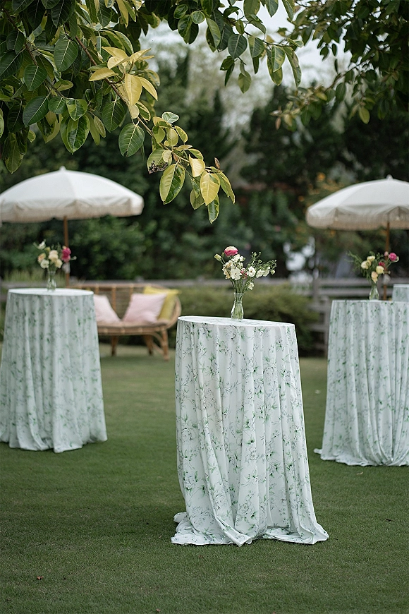 Cocktail hour decor with outdoor cocktail tables in green floral linens, bud vases and umbrellas, plus lounge sofa on a garden lawn