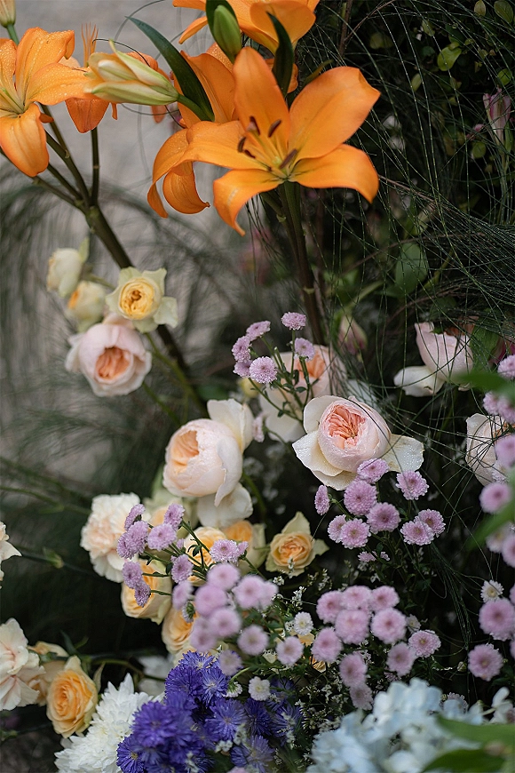 Wedding floral arrangement with orange lily wedding flowers, garden roses, lavender fillers, and greenery set against a neutral wall with foliage