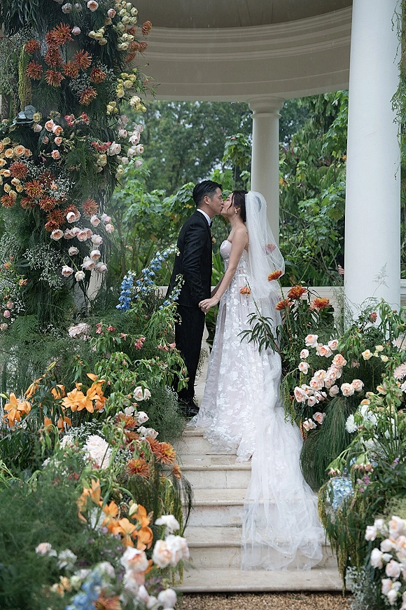 Wedding kiss portrait of bride and groom kissing, her long veil flowing by a floral arch of roses on white-column pavilion steps