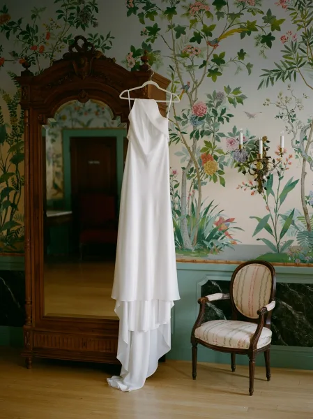 Wedding dress hanging on a dress hanger beside a mirror, framed by hand-painted floral wallpaper and a wall sconce with candles