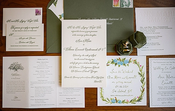 Wedding invitation suite flat lay with calligraphy cards, green envelopes, wax seal, ribbon and vintage stamps on a wood tabletop