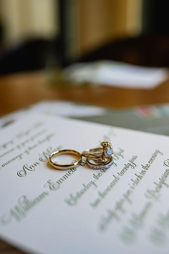 Wedding rings resting on a calligraphy invitation, featuring a diamond engagement ring and gold wedding band on a wood table
