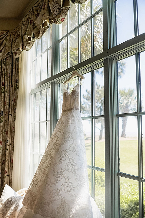Wedding dress with lace detailing and sweetheart neckline hanging on a window, jeweled belt sparkling in soft sunlight by trees