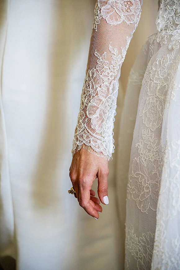 Wedding dress close-up showing lace sleeve wedding dress with floral appliqué on illusion tulle, bride’s hand with gold ring in soft light