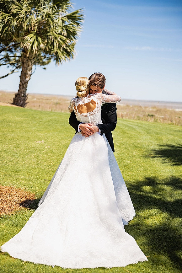 Wedding couple hug as bride in backless long-sleeve lace gown embraces groom in tuxedo on a green lawn with palm tree and blue sky